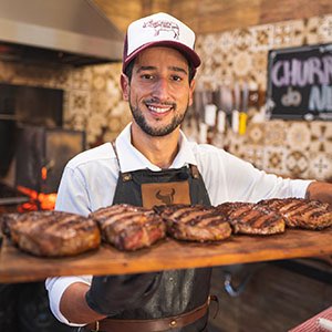 Netão sorrindo segurando uma tábua de carnes assadas